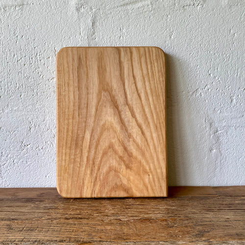 A rectangular Ash wood cutting board with rounded corners, standing upright on a wooden table against a textured white wall.