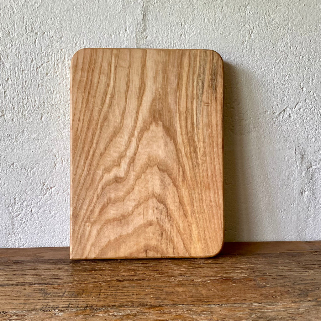 A rectangular Ash wood cutting board with rounded corners, standing upright on a wooden table against a textured white wall.