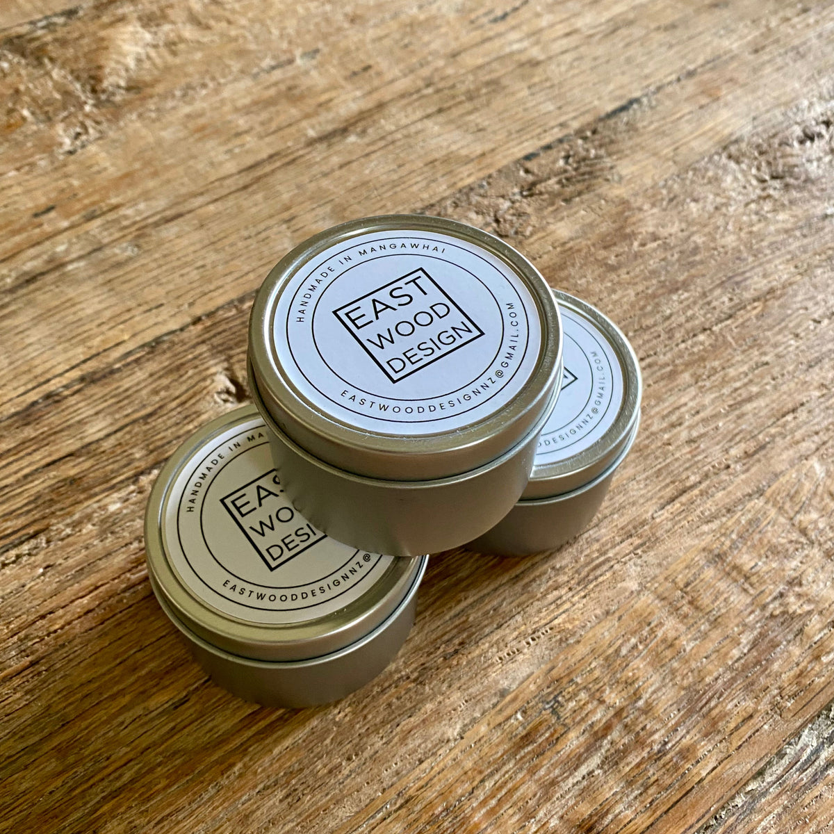 Three small round metal tins of beeswax mineral oil conditioner with white lids printed with the brand name 'East Wood Design' arranged on a rustic wooden tabletop.