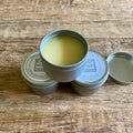 Three small round metal tins of beeswax mineral oil wax on a rustic wooden surface, with one tin open to show the pale yellow product inside and the other lids closed. The lid label reads East Wood Design.