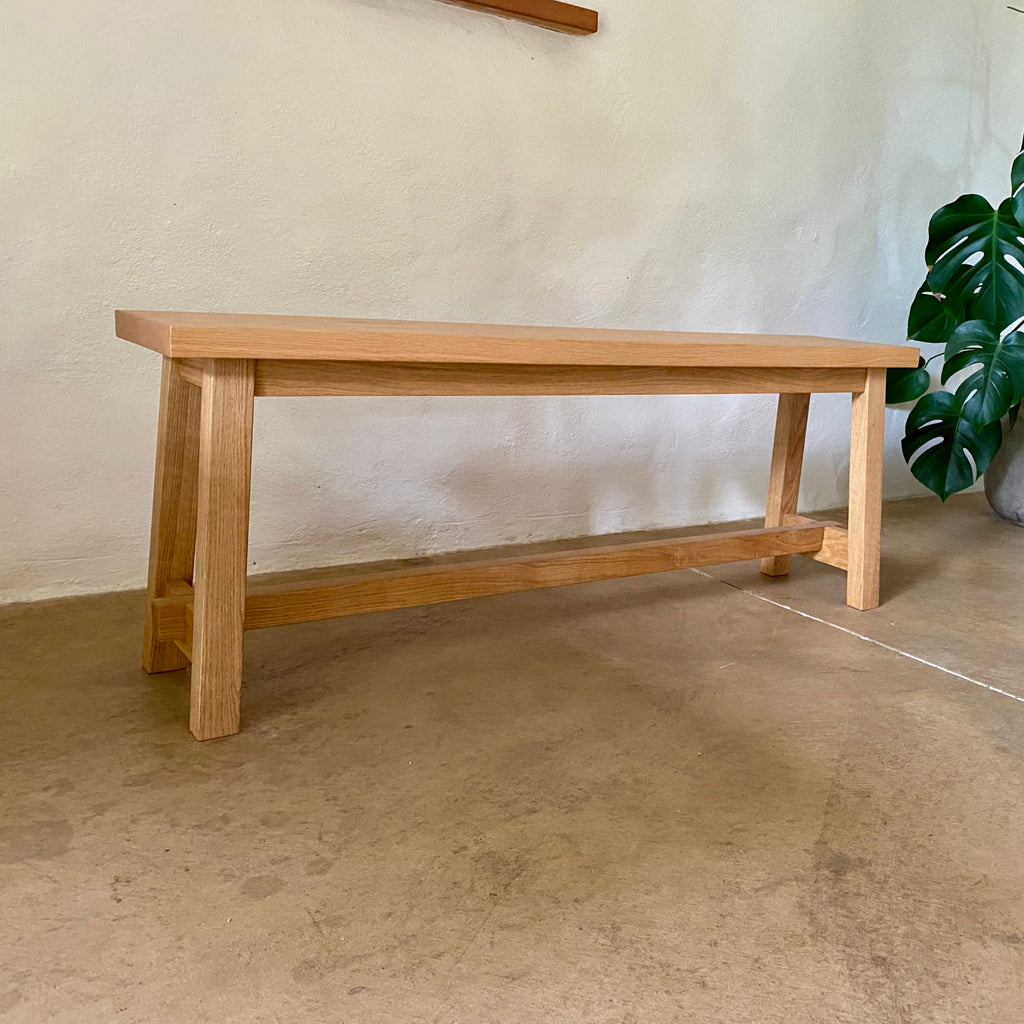 A light natural wood bench with a rectangular top, four legs, and a lower support rail, photographed indoors against a textured off-white wall with a concrete floor and a green plant partially visible on the right.