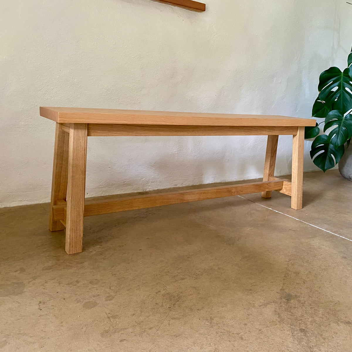 A light natural wood bench with a rectangular top, four legs, and a lower support rail, photographed indoors against a textured off-white wall with a concrete floor and a green plant partially visible on the right.