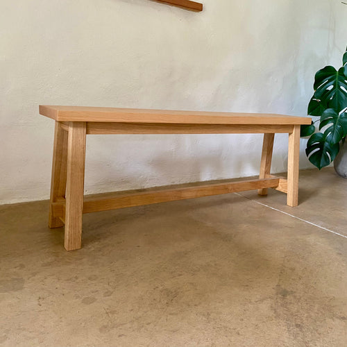 A light natural wood bench with a rectangular top, four legs, and a lower support rail, photographed indoors against a textured off-white wall with a concrete floor and a green plant partially visible on the right.