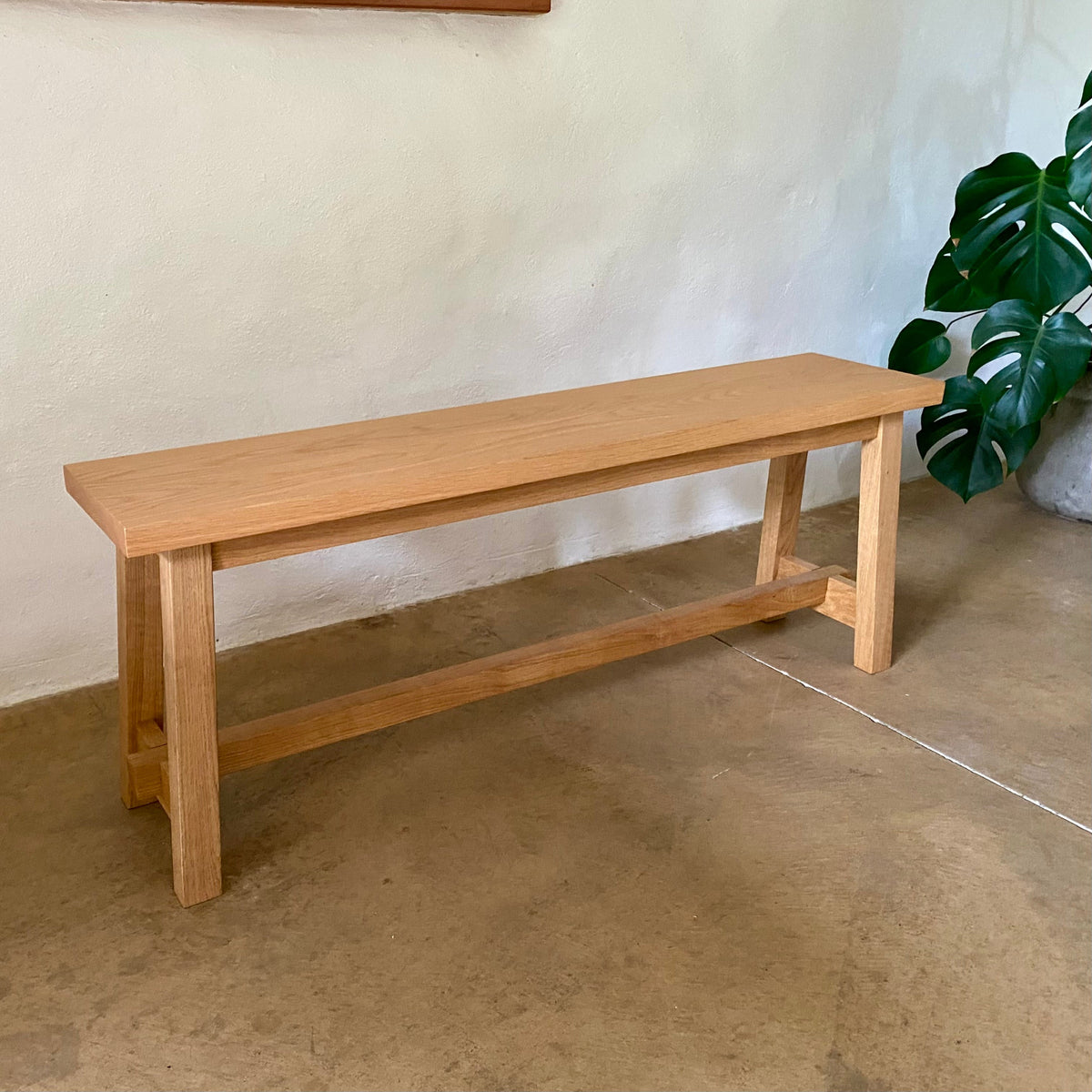 A light natural wood bench with a rectangular seat and open lower support rail, placed on a concrete floor against a textured off-white wall with a green plant in the background.