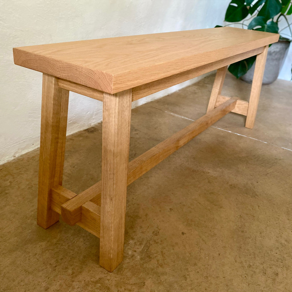 A light natural oak wooden bench with a thick rectangular seat and trestle-style legs, photographed in a real indoor setting with a stucco wall, concrete floor, and a potted plant in the background.
