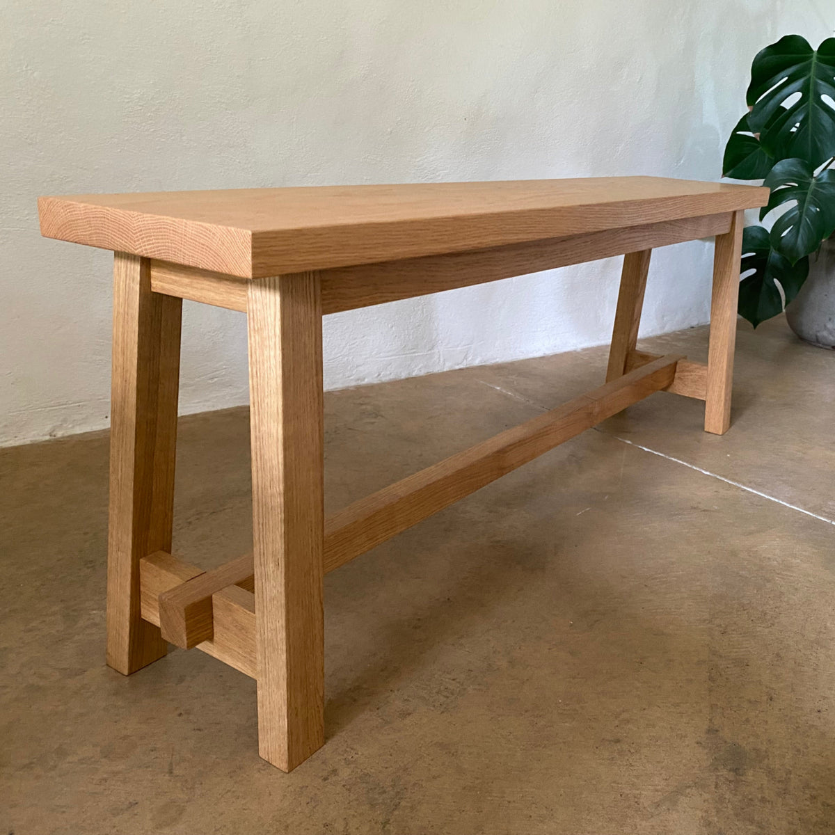 A light natural oak wood bench with a rectangular top, four legs, and a lower support rail, shown in a minimalist room with a concrete floor and a potted plant in the background.