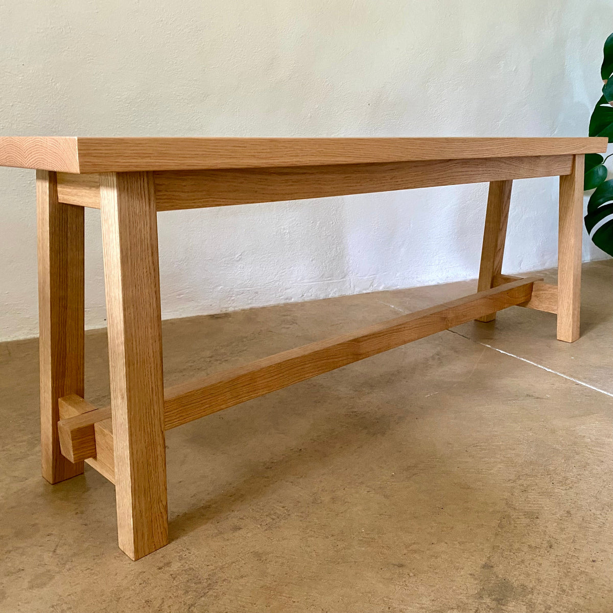 A light natural oak wooden bench with a thick rectangular top, four sturdy legs, and a lower support rail, shown in a minimalist indoor setting with a textured wall and concrete floor.