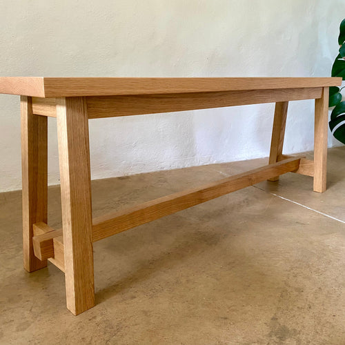 A light natural oak wooden bench with a thick rectangular top, four sturdy legs, and a lower support rail, shown in a minimalist indoor setting with a textured wall and concrete floor.