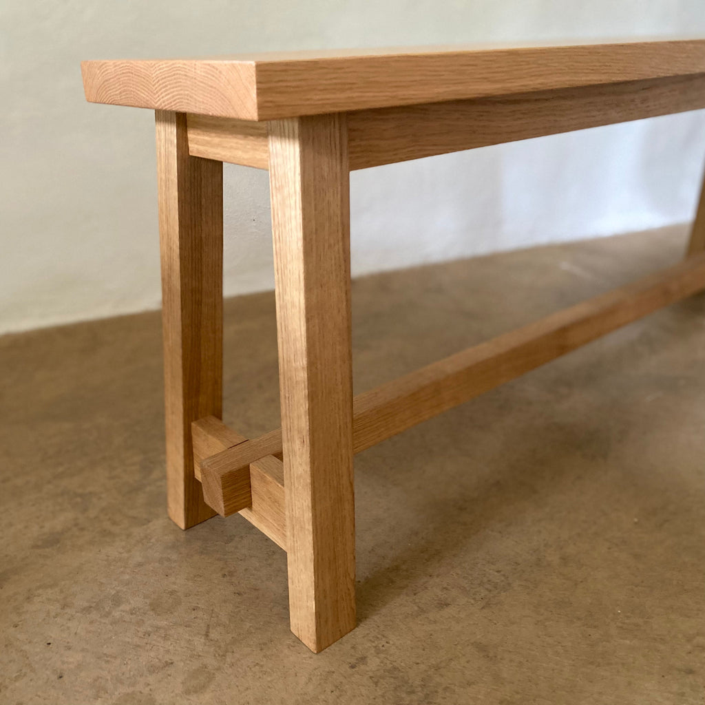 A close-up of a light oak wood bench showing the front legs, trestle-style joinery, and a thin wooden apron running beneath the seat, set against a neutral concrete floor and wall.