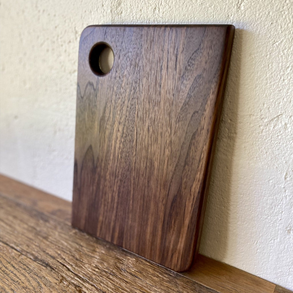 Small black walnut chopping board with finger hole leaning against white wall – East Wood Design