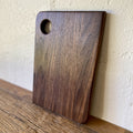 Small black walnut chopping board with finger hole leaning against white wall – East Wood Design