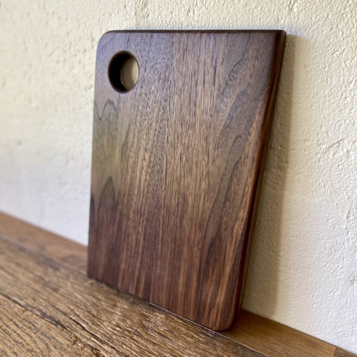 Small black walnut chopping board with finger hole leaning against white wall – East Wood Design