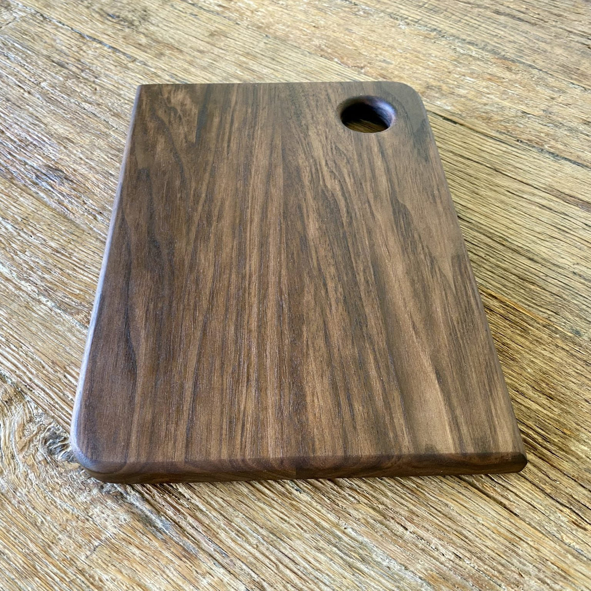 Small black walnut chopping board with finger hole, flat lay on rustic timber surface – East Wood Design