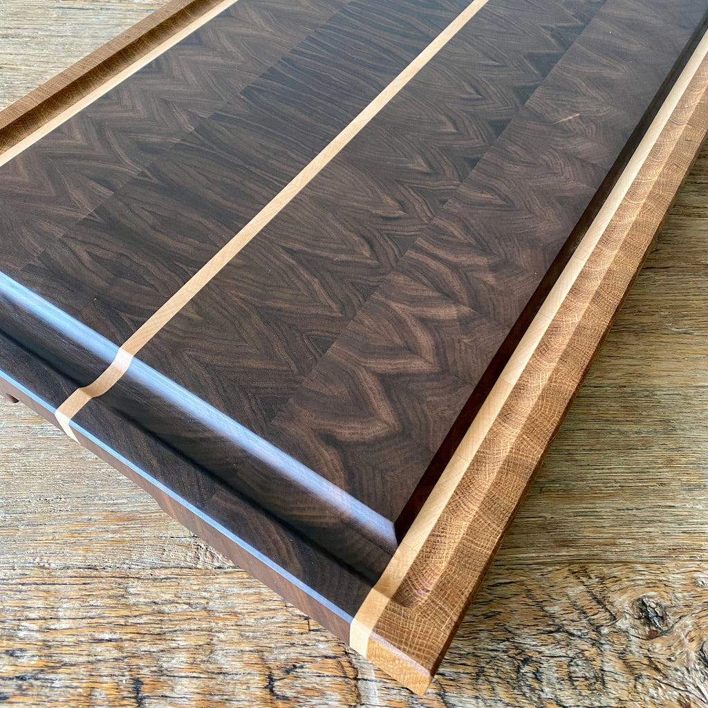 A close up image of rectangular Black Walnut end grain chopping board with Maple and White Oak stripes on the sides and a Maple stripe in the middle, It has a juice groove around the boarder and it sits on a rust table