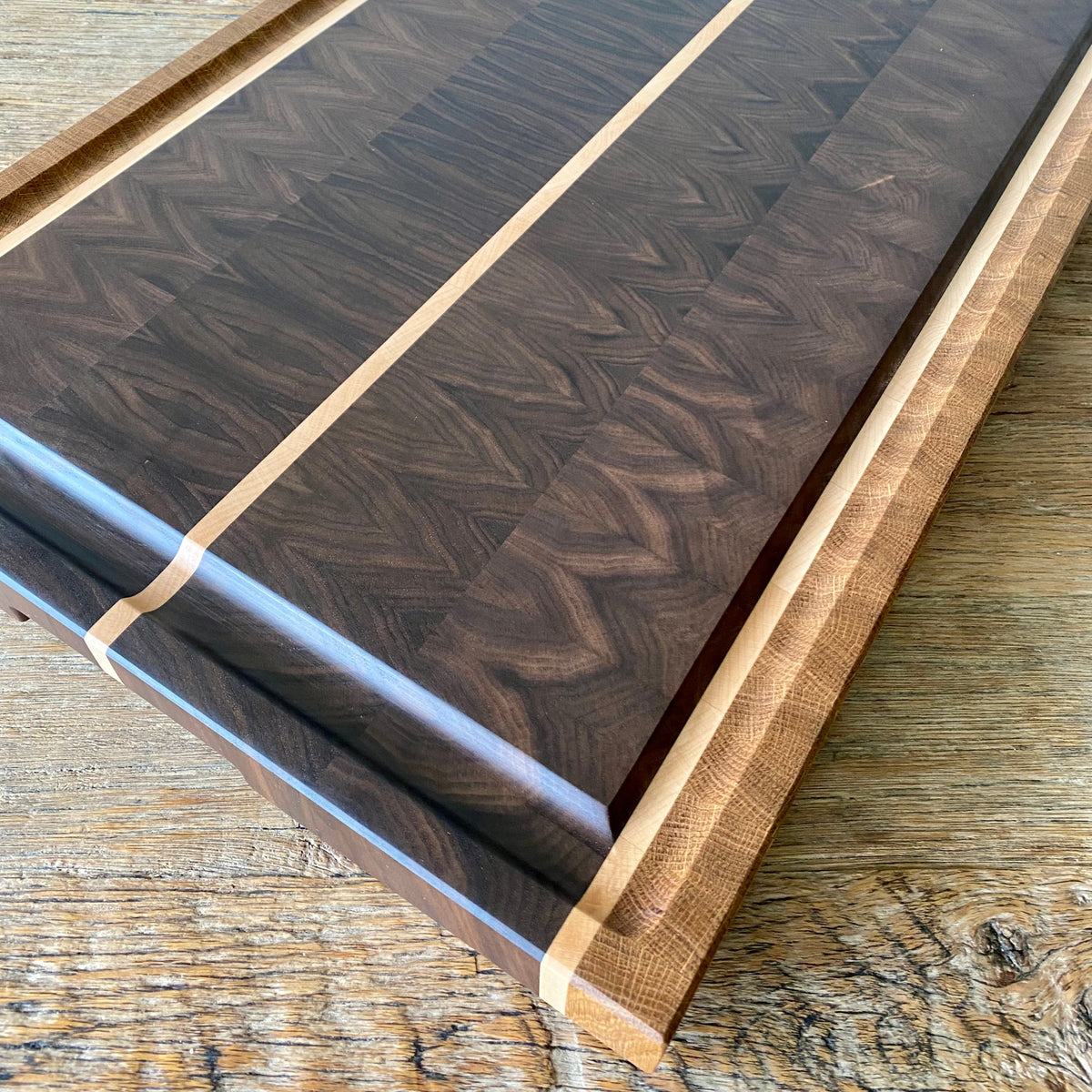 A close up image of rectangular Black Walnut end grain chopping board with Maple and White Oak stripes on the sides and a Maple stripe in the middle, It has a juice groove around the boarder and it sits on a rust table