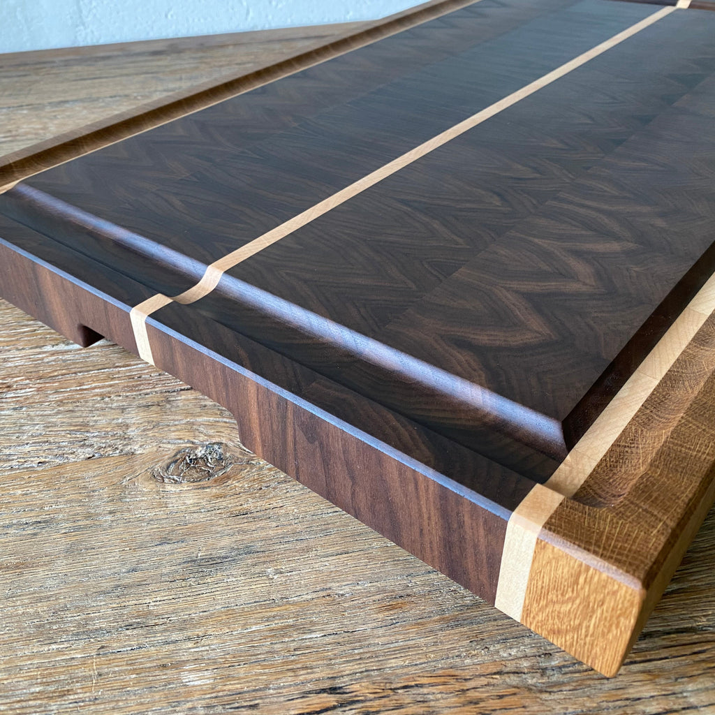 A close up of a rectangular Black Walnut end grain chopping board with Maple and White Oak stripes on the sides and a Maple stripe in the middle, It has a juice groove around the boarder and a cut out handle it sits on a rust table