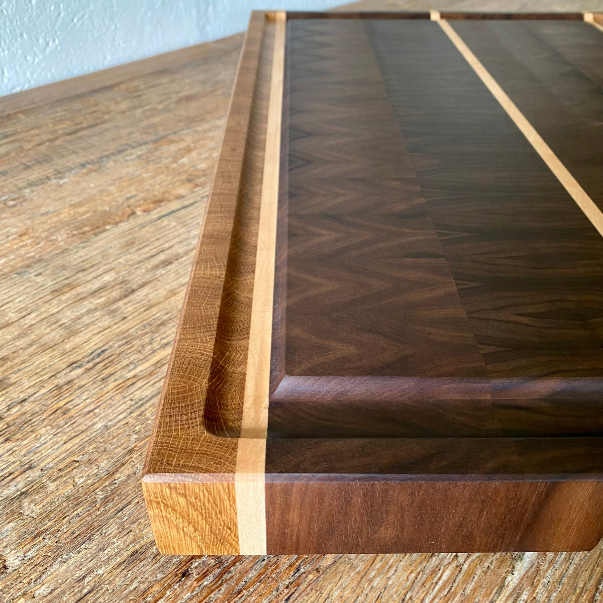 A close-up of a rectangular Black Walnut chopping board with a dark brown chevron-patterned center and light natural wood borders, resting on a rustic wooden tabletop.
