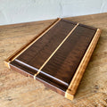 A rectangular Black Walnut end grain chopping board with Maple and White Oak stripes on the sides and a Maple stripe in the middle, It has a juice groove around the boarder and it sits on a rust table with a white wall behind