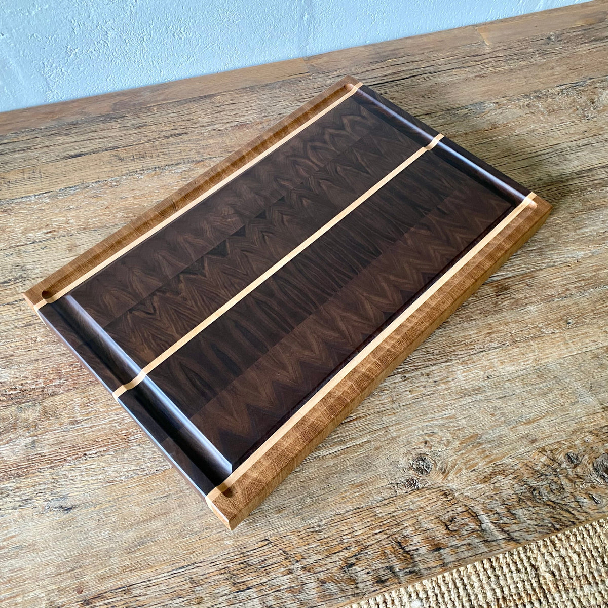 A rectangular Black Walnut end grain chopping board with Maple and White Oak stripes on the sides and a Maple stripe in the middle, It has a juice groove around the boarder and it sits on a rust table