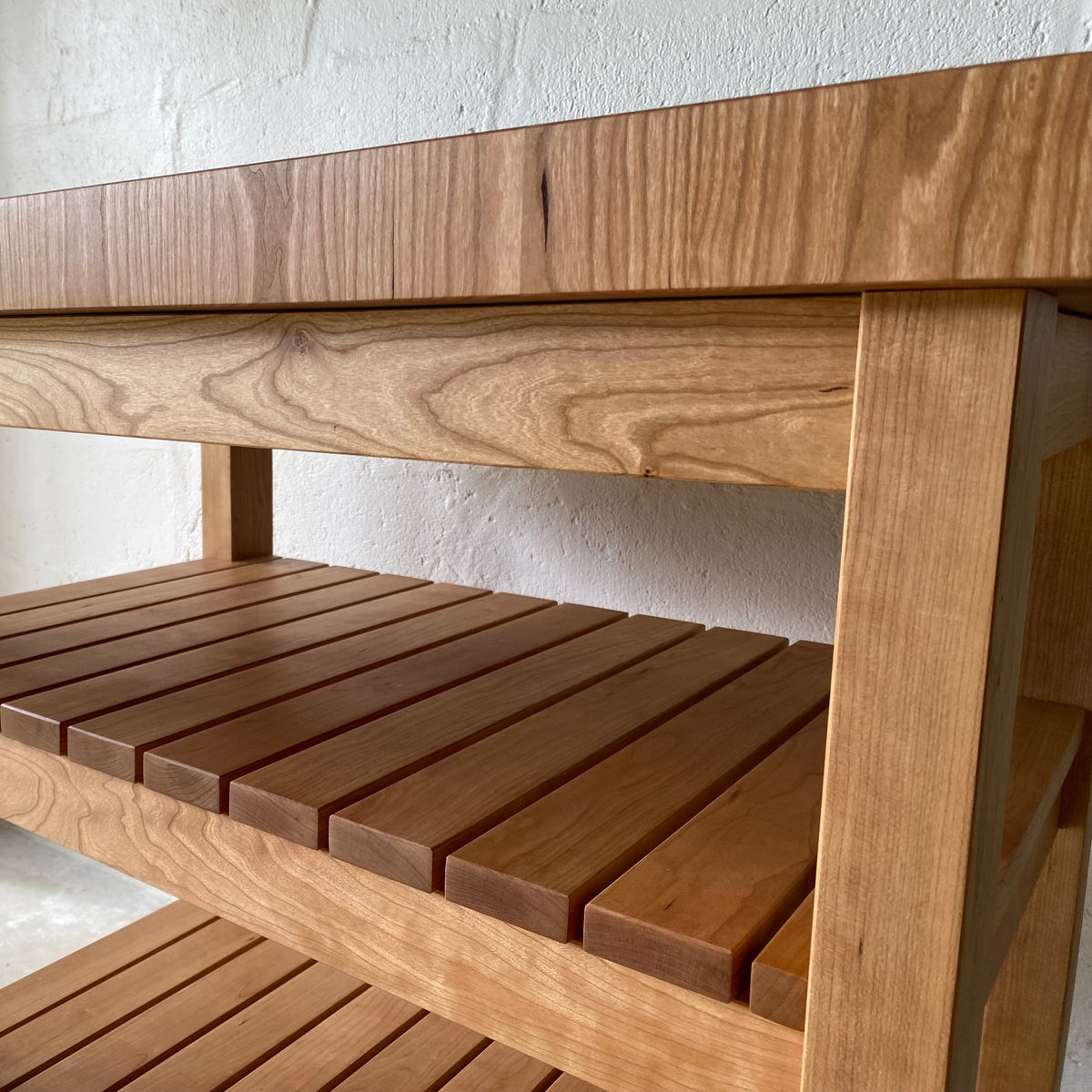 A close-up of a wooden butcher island made from cherry with a warm medium-brown finish, showing the wood grain on the top surface and slatted shelves, set against a light texture