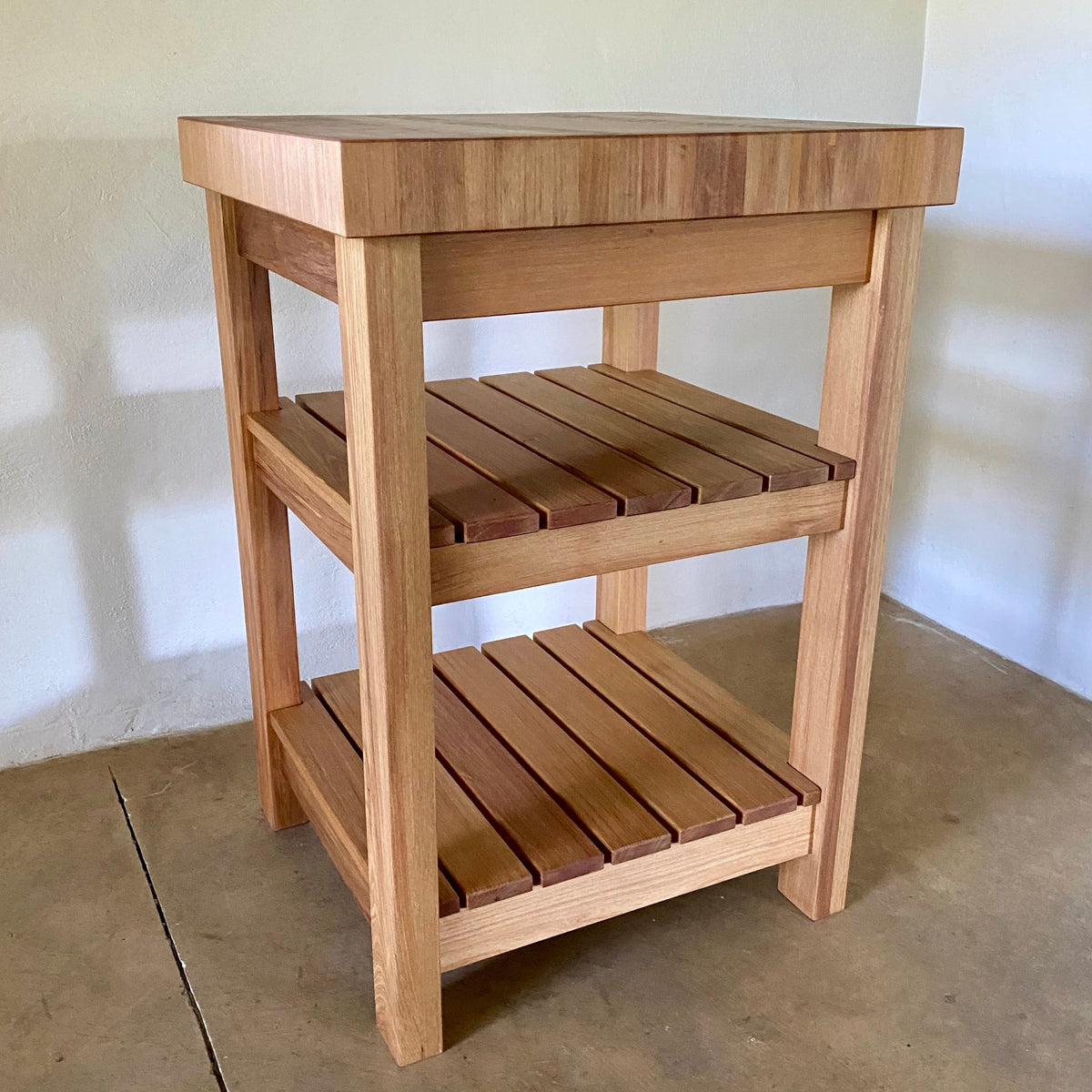 A end grain butcher island, made from pacific rimu with a middle slatted shelf, and a bottom slatted shelf, photographed in a room corner.