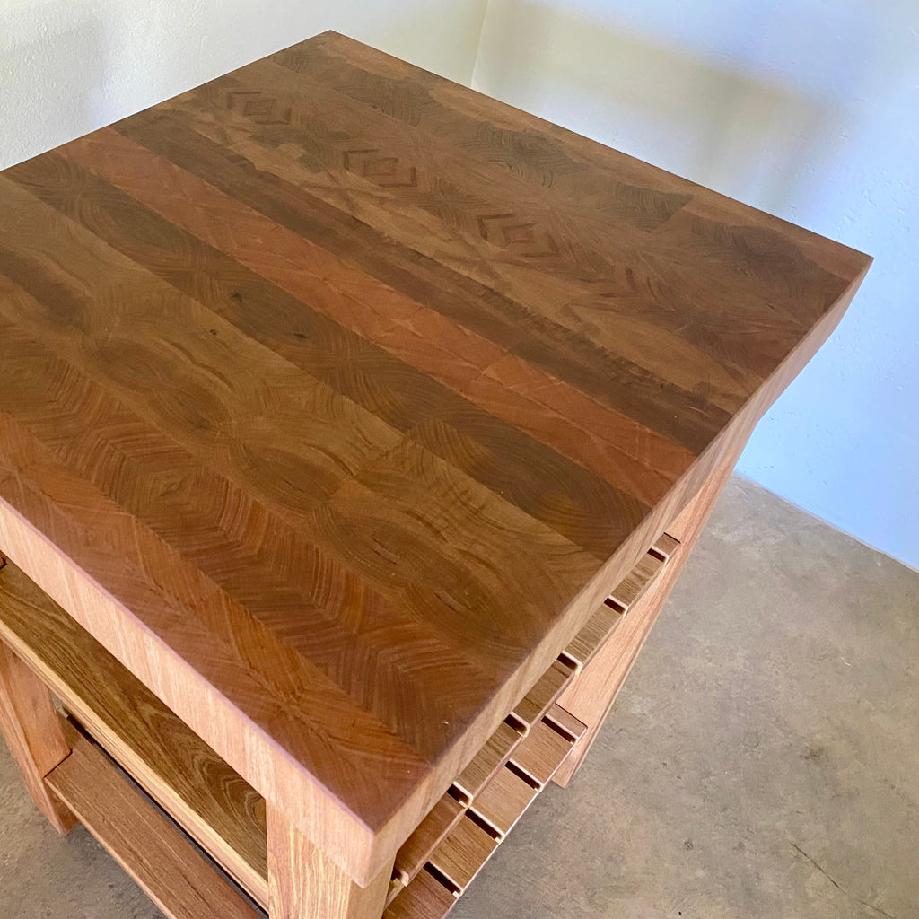 A end grain butcher island, made from pacific rimu, showing the thick end grain pattern top with a middle slatted shelf, and a bottom slatted shelf, photographed in a room corner.