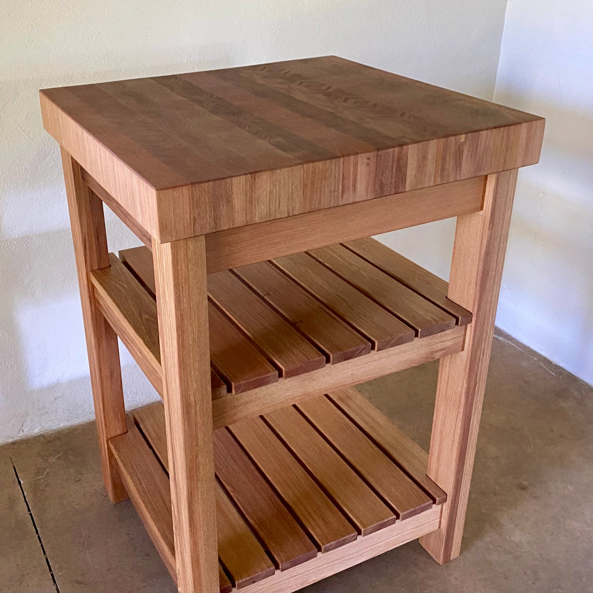 A end grain butcher island, made from pacific rimu with a middle slatted shelf, and a bottom slatted shelf, photographed in a room corner. showing thick end grain patterned top