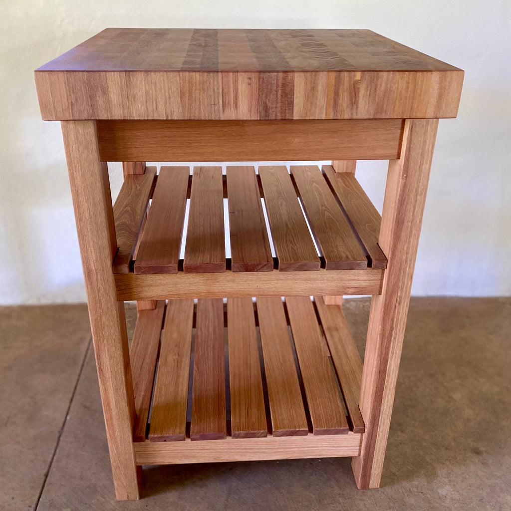 A end grain butcher island, made from pacific rimu with a middle slatted shelf, and a bottom slatted shelf, photographed straight on with a plain back ground and concrete floor 
