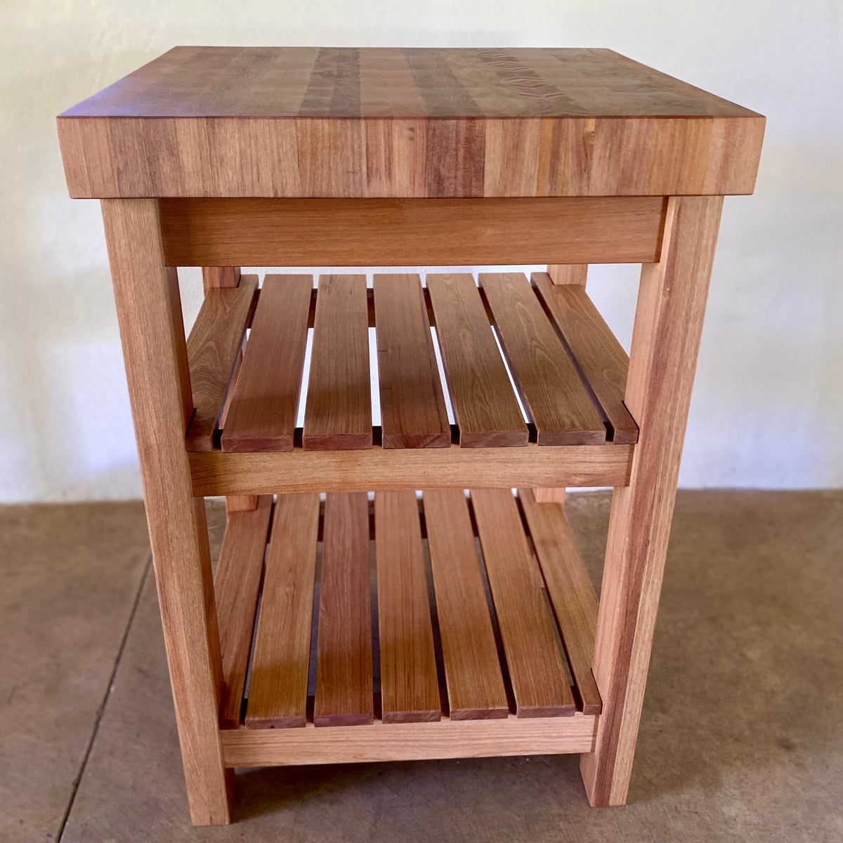 A end grain butcher island, made from pacific rimu with a middle slatted shelf, and a bottom slatted shelf, photographed straight on with a plain back ground and concrete floor 