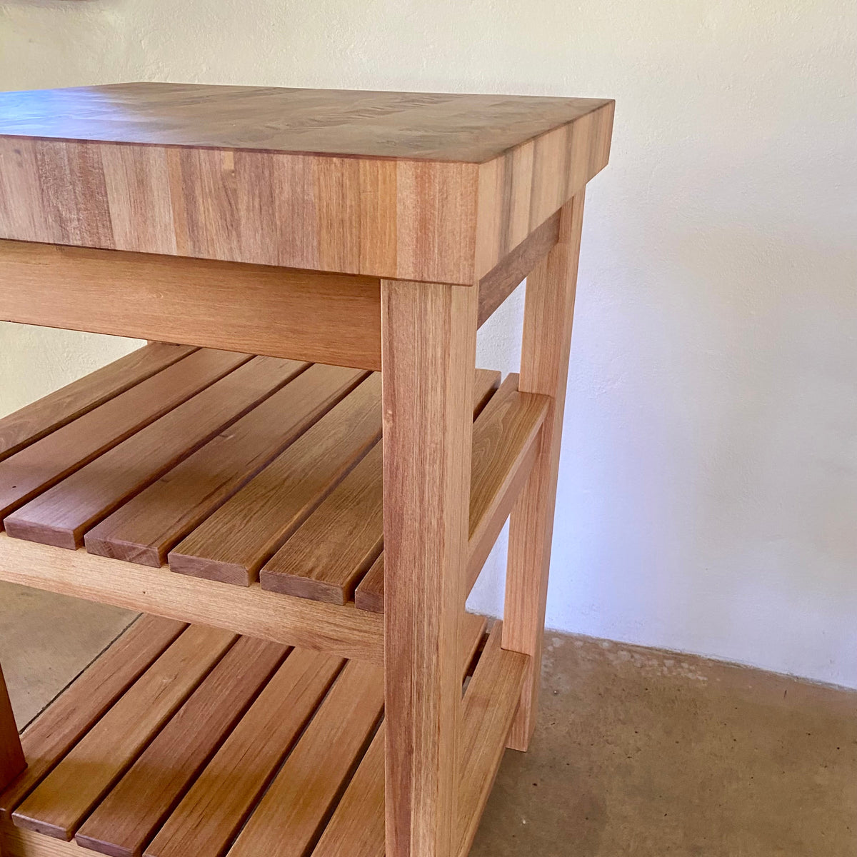 A end grain butcher island, made from pacific rimu with a middle slatted shelf, and a bottom slatted shelf,  with a thick rectangular top and open side supports, shown in a real indoor setting with a plain wall and concrete floor.