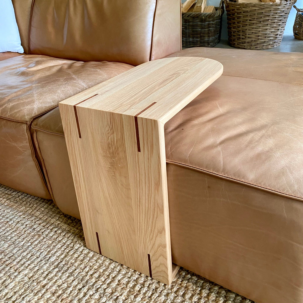 A light White Ash wooden C side table with a curved top and minimalist design, shown leaning against a tan leather sectional sofa in a living room setting.