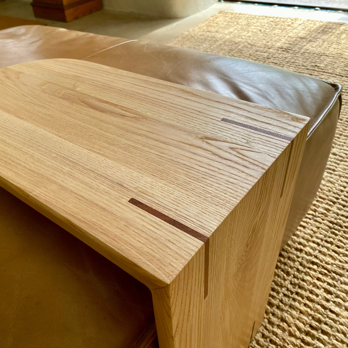 Close-up of a light White Ash wooden C coffee table with visible grain and inlay details, positioned next to a brown leather sofa on a woven beige rug.