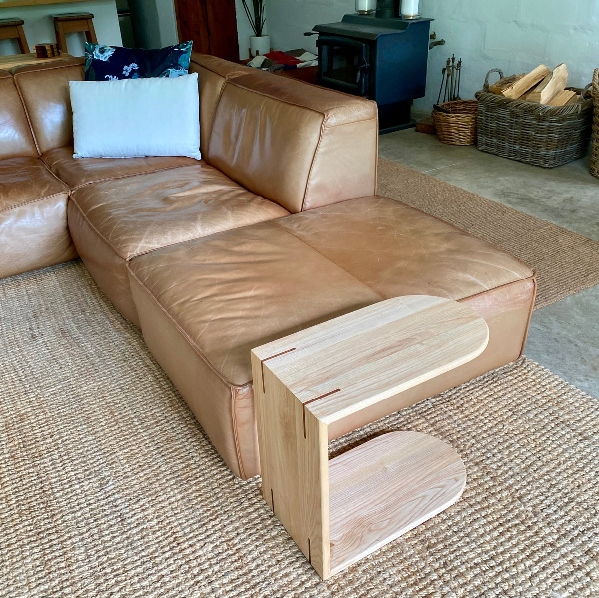 A light natural White Ash wooden C side table placed in front of a tan leather sectional sofa in a real living room setting with a woven rug, wood stove, and baskets of firewood