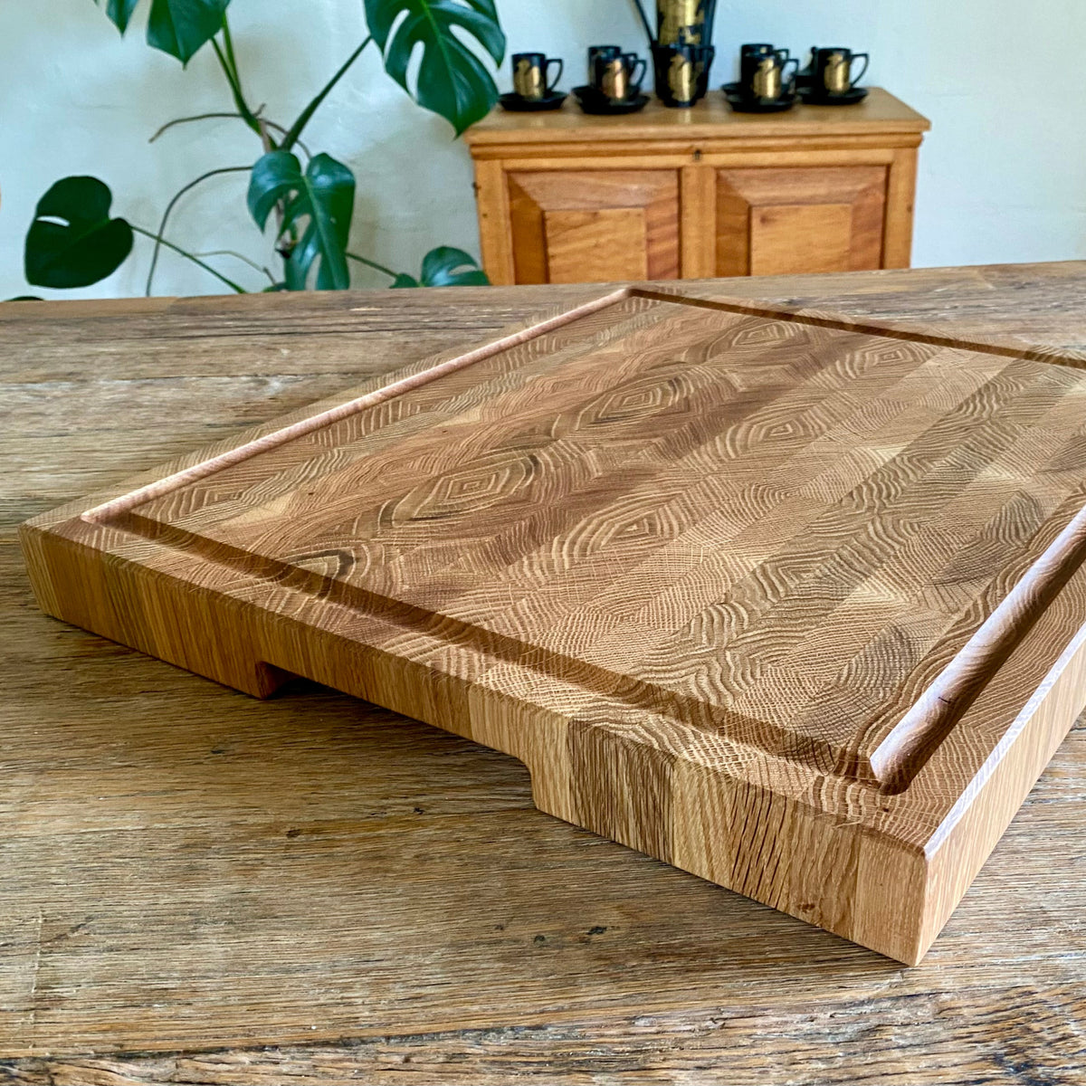 A thick, square handcrafted white oak end grain cutting board with a prominent natural wood grain pattern, resting on a rustic wooden table in a home setting.