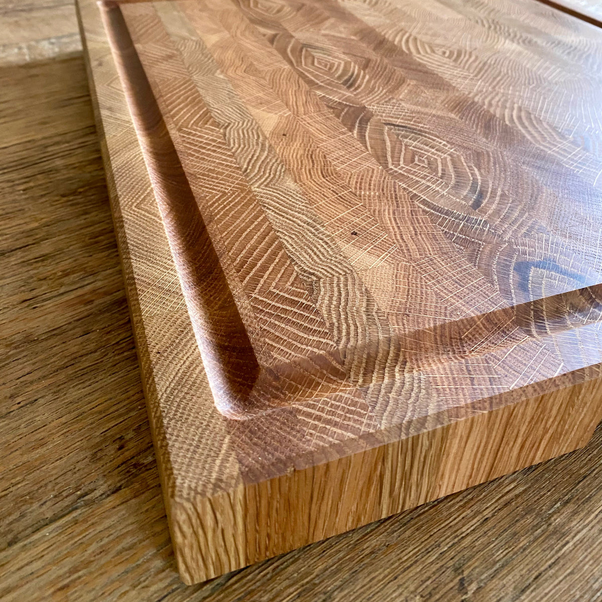 A thick, white oak wooden end grain cutting board with a juice groove featuring a rich grain pattern, resting on a wooden table in a home setting.