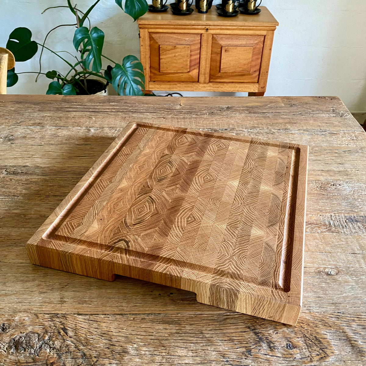 A white oak end grain chopping board with distinctive grain pattern, juice grove and cut out handle on a rustic table