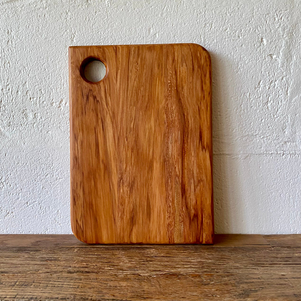 A rectangular heart rimu wooden cutting board with rounded corners and a circular handle cutout in the upper left, standing upright on a wooden surface against a textured white wall.