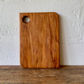 A rectangular heart rimu wooden cutting board with rounded corners and a circular handle cutout in the upper left, standing upright on a wooden surface against a textured white wall.