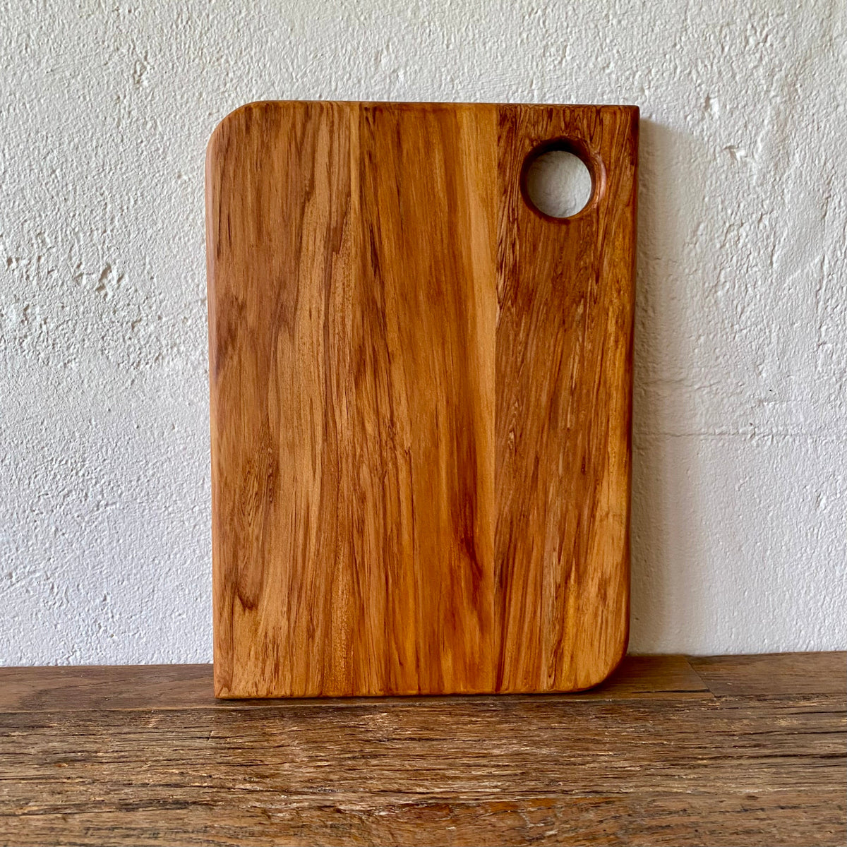 A rectangular heart rimuwooden cutting board with rounded corners and a circular handle cutout in the top right, standing upright on a wooden surface against a textured off-white wall.