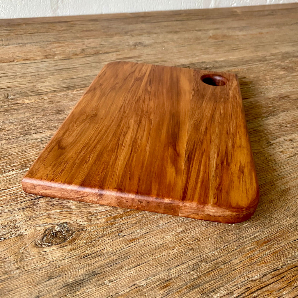 A rectangular heart Rimu wooden cutting board with rounded corners and a small oval handle cutout in the upper right, shown on a rustic wooden tabletop. 