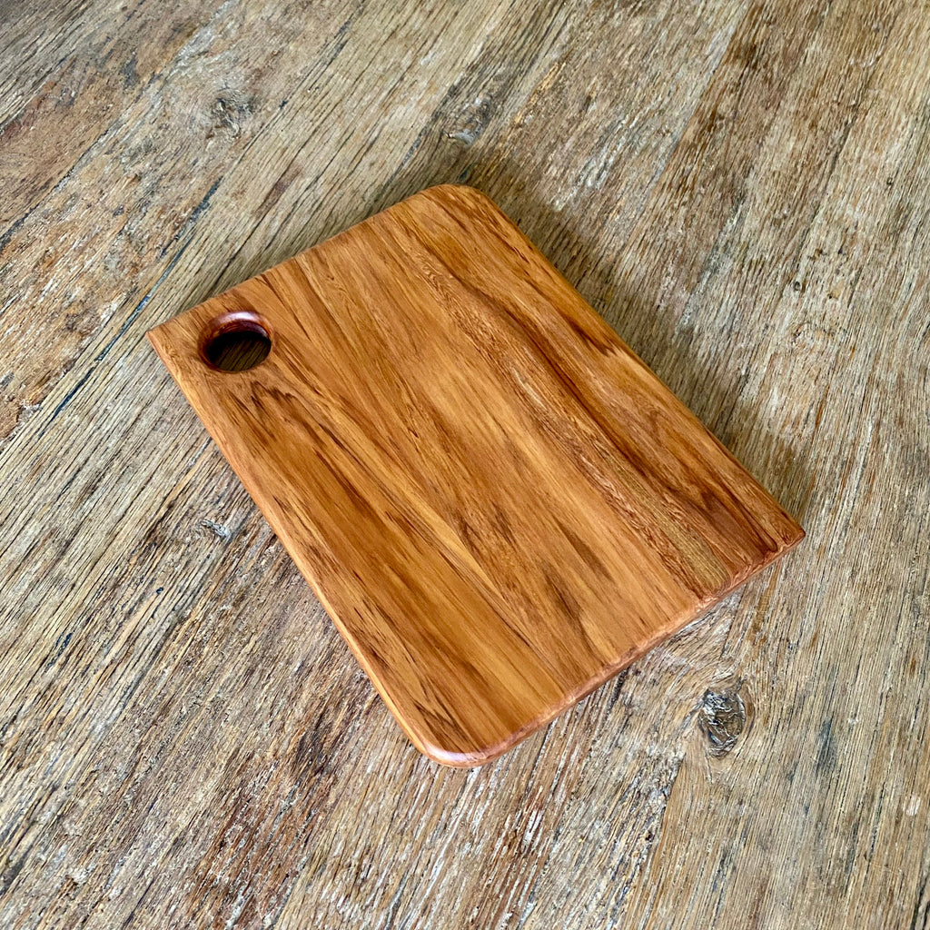 A rectangular heart Rimu wooden cutting board with rounded corners and a small oval hole near one corner, shown on a rustic wood surface.