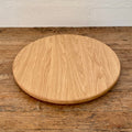 A round natural white oak lazy Susan, turntable with visible wood grain, displayed on a rustic wooden tabletop against a white wall.