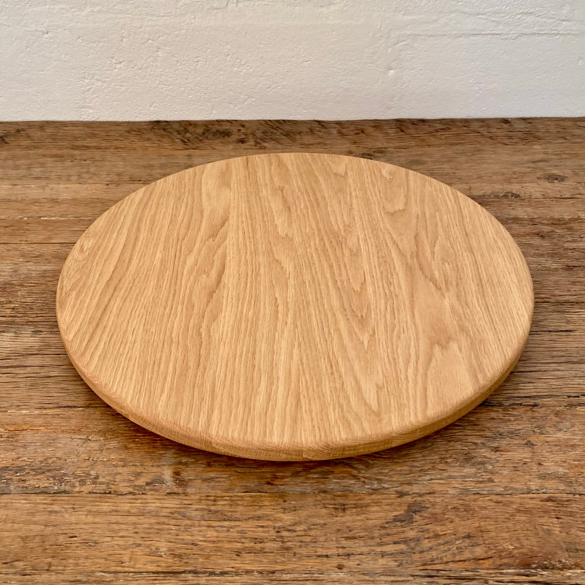 A round natural white oak lazy Susan, turntable with visible wood grain, displayed on a rustic wooden tabletop against a white wall.