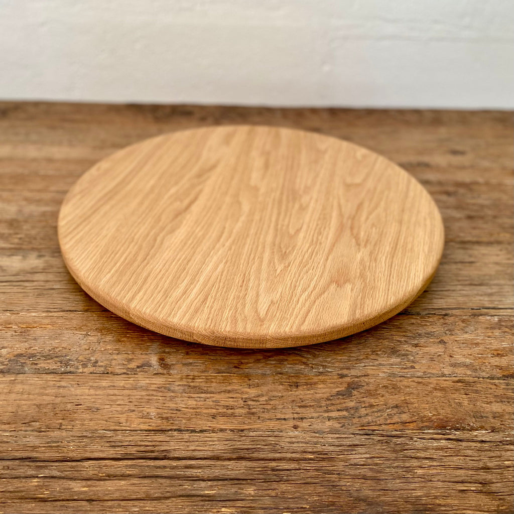 A round white oak lazy Susan, turntable with a natural oak finish, placed on a rustic wooden table against a white brick wall.