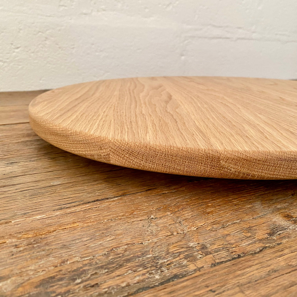 A round white oak lazy Susan, turntable with a natural oak grain, resting on a rustic wooden table against a white brick wall.