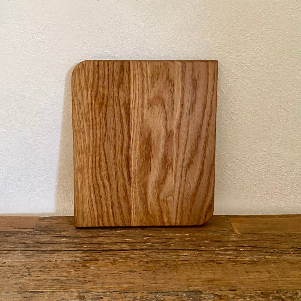 A square white oak cutting board with rounded corners and a prominent vertical grain pattern, standing upright on a rustic wooden surface against a textured off-white wall.
