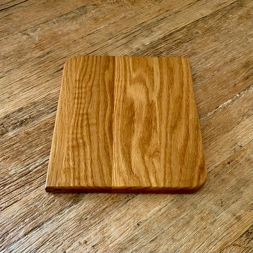 A square white oak wood cutting board with rounded corners and a smooth, polished finish, placed on a rustic wooden table.