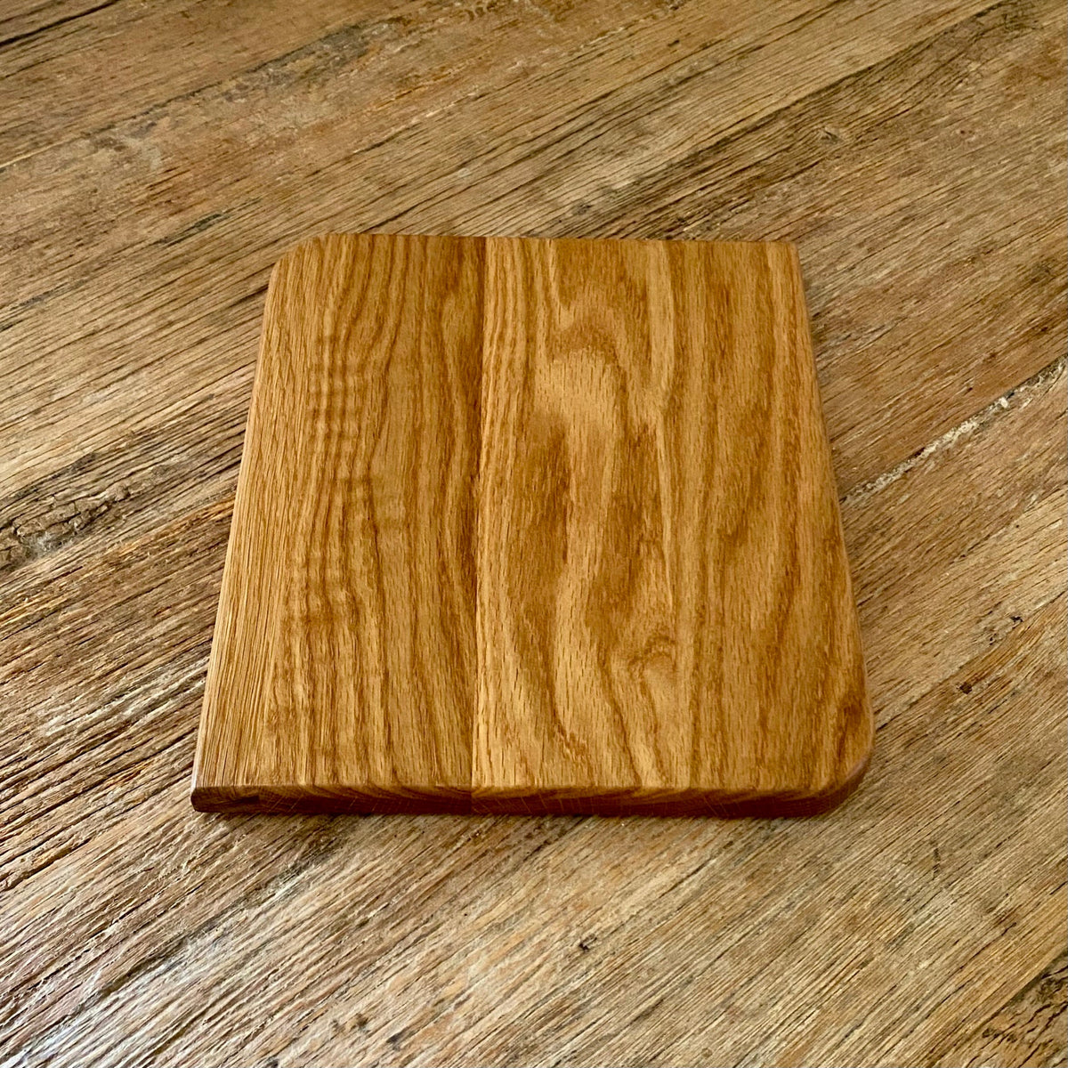 A square white oak wood cutting board with rounded corners and a smooth, polished finish, placed on a rustic wooden table.