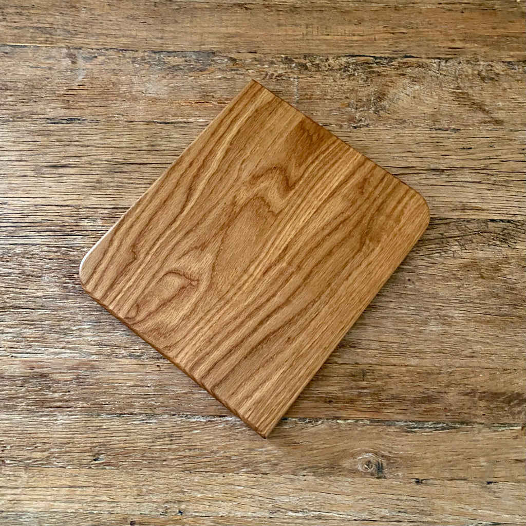 A square white oak wooden cutting board with rounded corners, displayed diagonally on a rustic wooden tabletop.