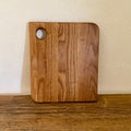 A square oak wooden cutting board with rounded corners and a circular handle cutout in the upper left, standing upright on a wooden surface against a textured off-white wall.